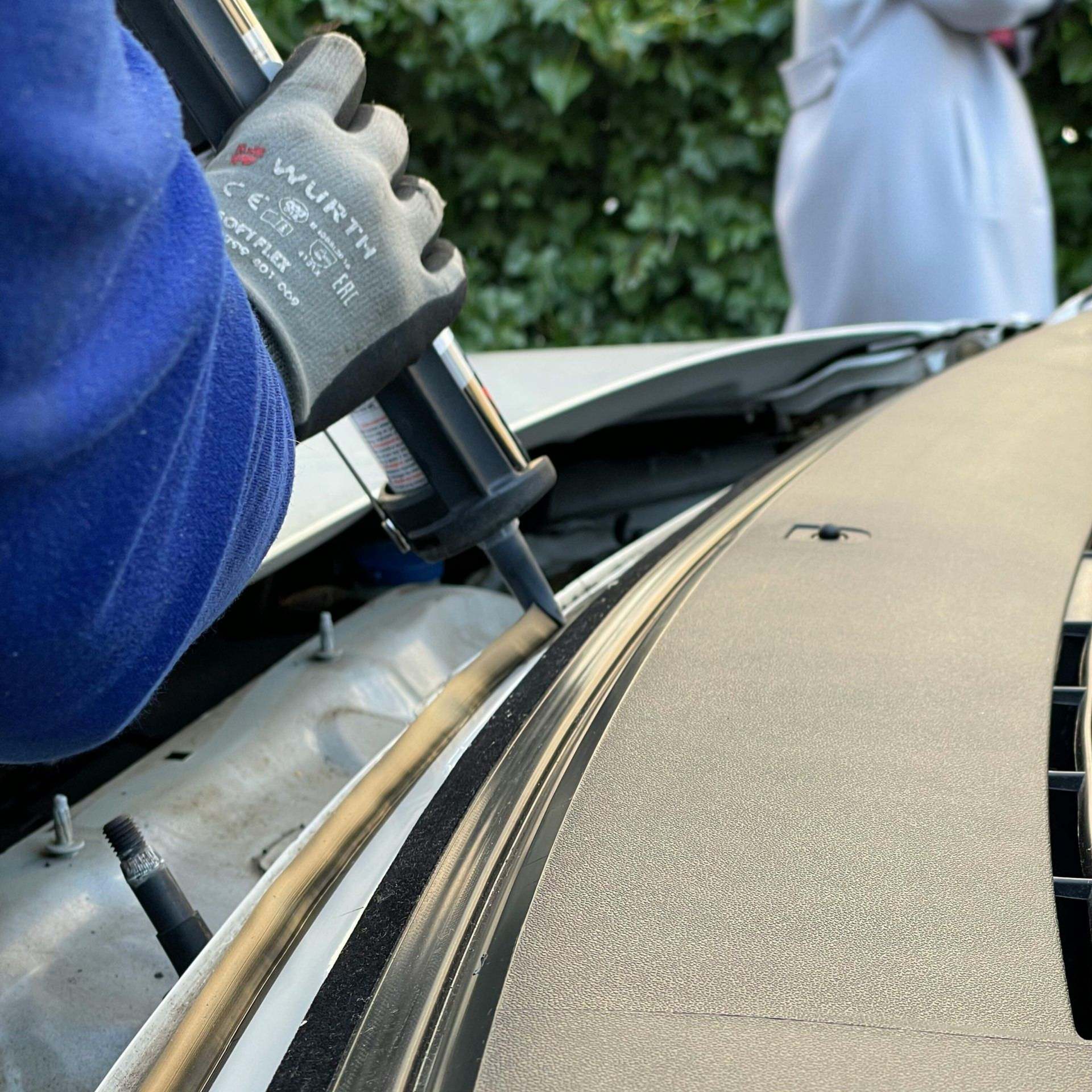 Person applying sealant to a car windshield seam with a caulking gun outdoors.