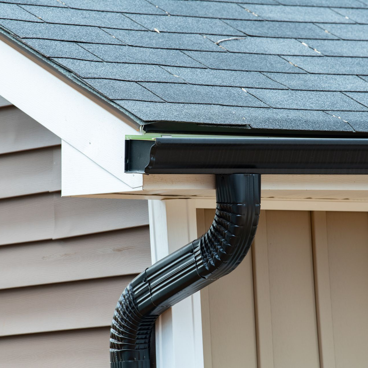 Black rain gutter attached to a house with brown siding and a gray shingled roof.
