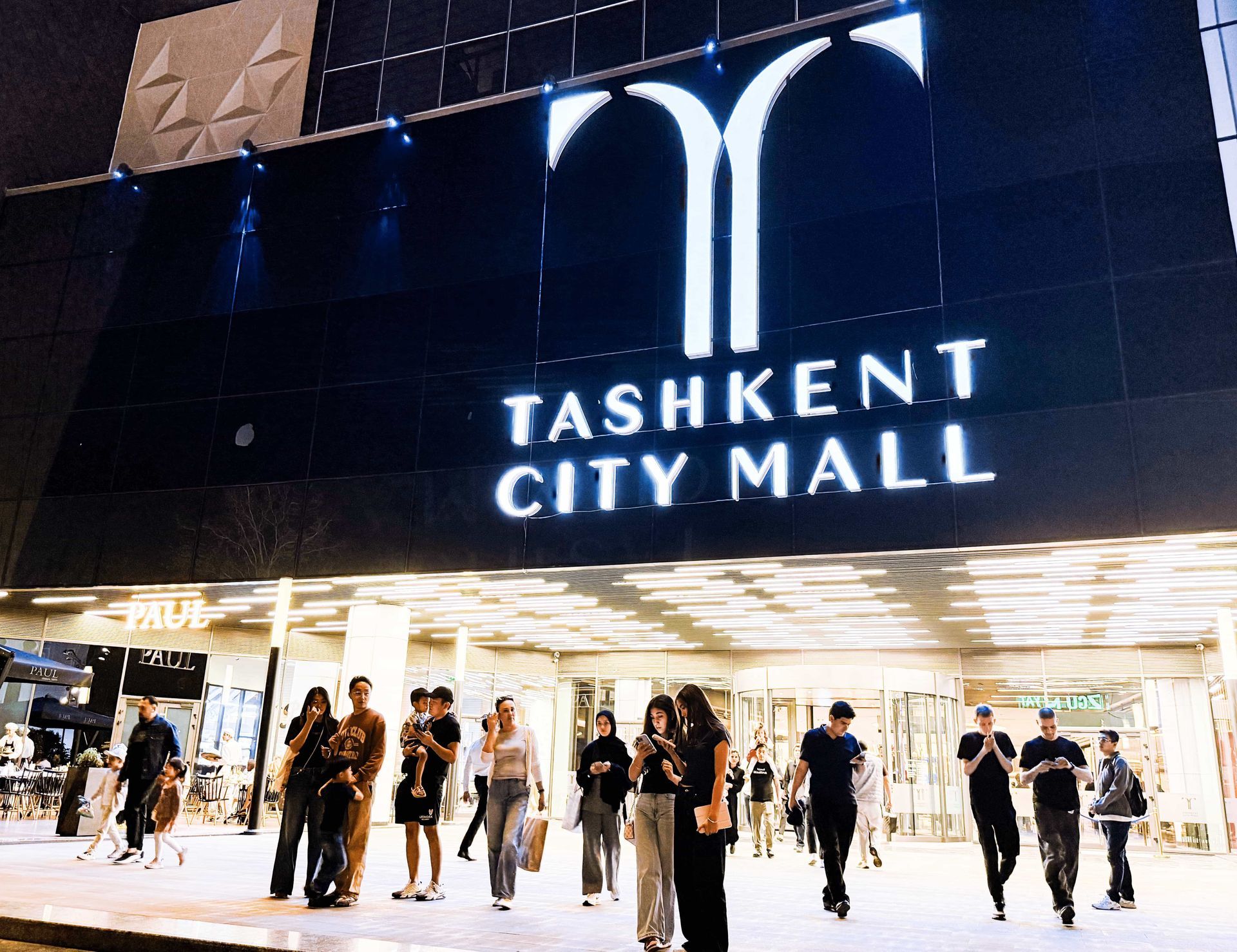 Entrance of Tashkent City Mall at night, with bright white lights and people walking in front of the sign