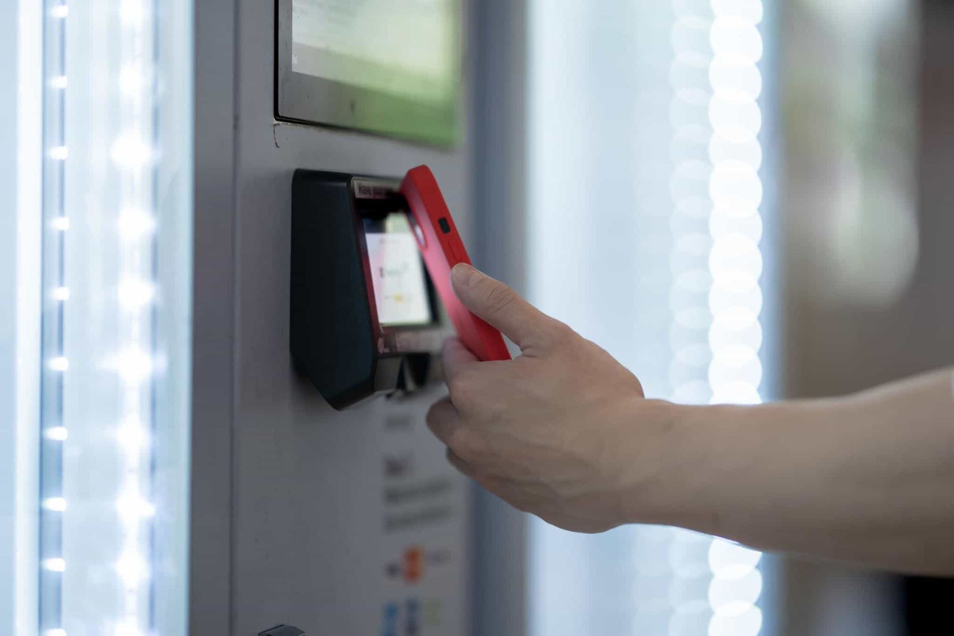 A hand holds a red smartphone up to a digital scanner on a kiosk to make a contactless payment.