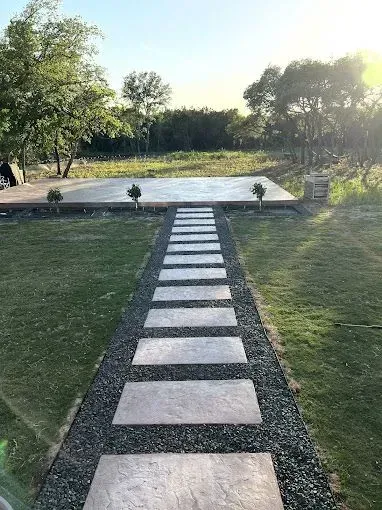 A stone walkway leading to a concrete platform in the middle of a lush green field.