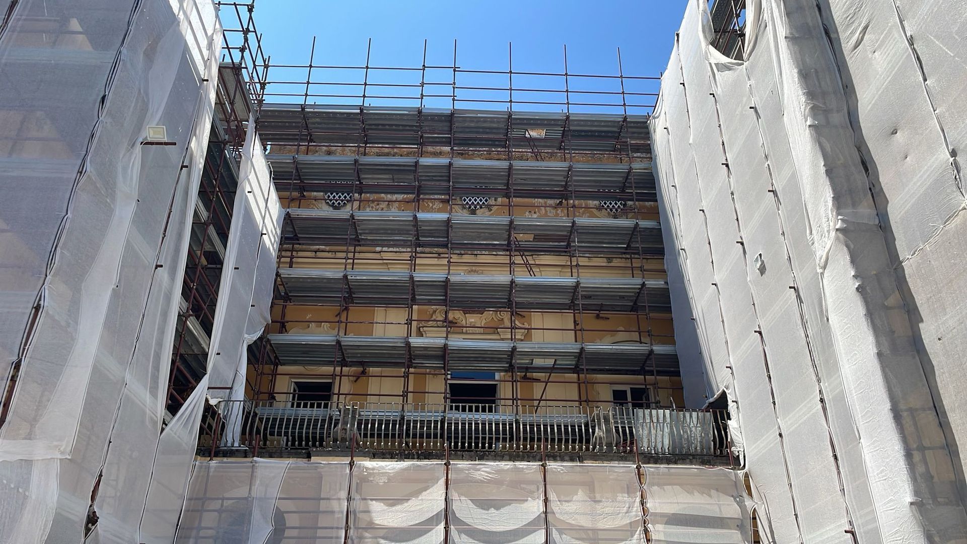 Facciata di un edificio in fase di ristrutturazione, coperta da impalcature e reti protettive. Cielo soleggiato e luminoso.