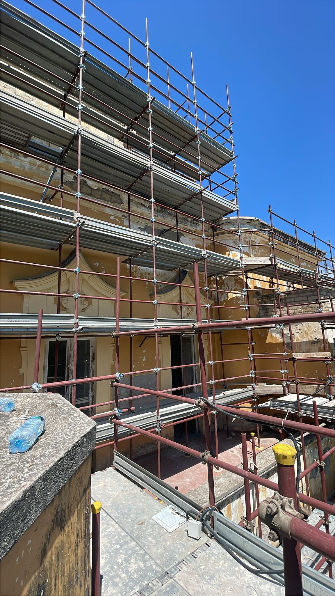 Un'impalcatura circonda l'esterno deteriorato di un edificio durante un apparente lavoro di restauro; cielo soleggiato in alto.