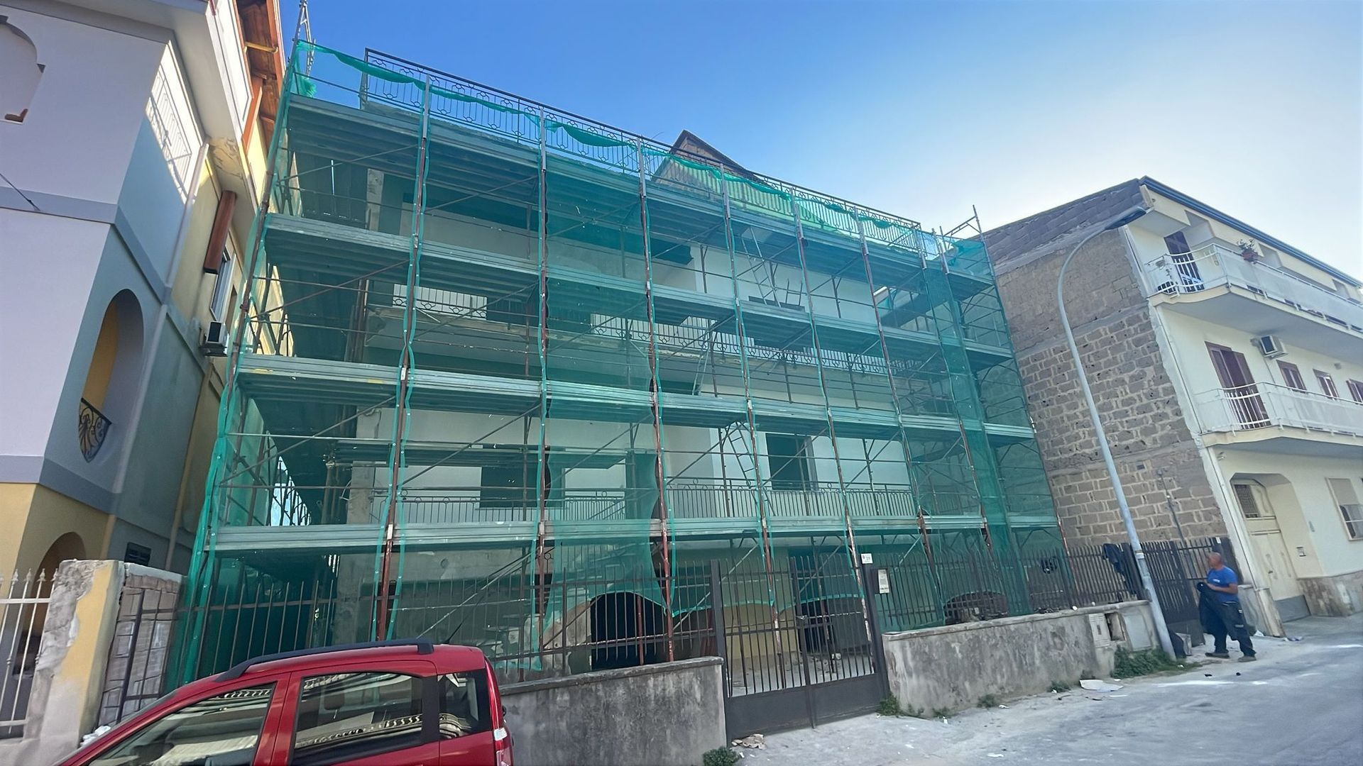 Edificio in costruzione, impalcature coperte da rete di sicurezza verde. Auto rossa in primo piano.