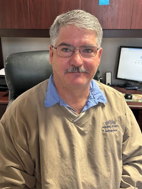Man in glasses, tan sweater, blue shirt collar, seated at desk.