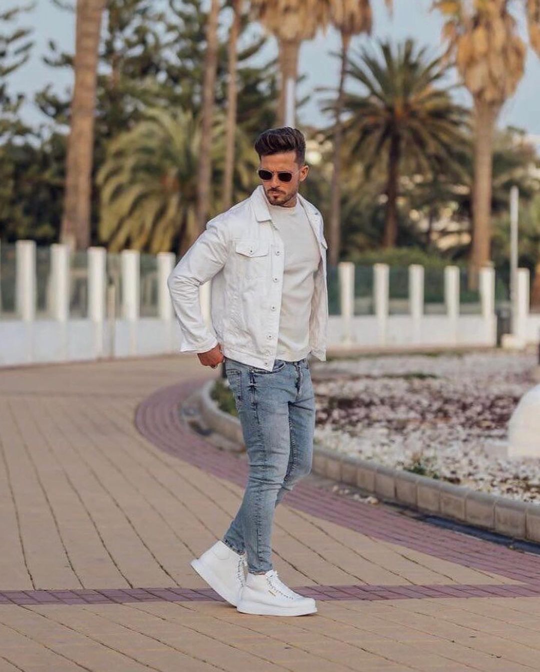 Un hombre que lleva una chaqueta vaquera blanca y pantalones vaqueros está parado en una acera.