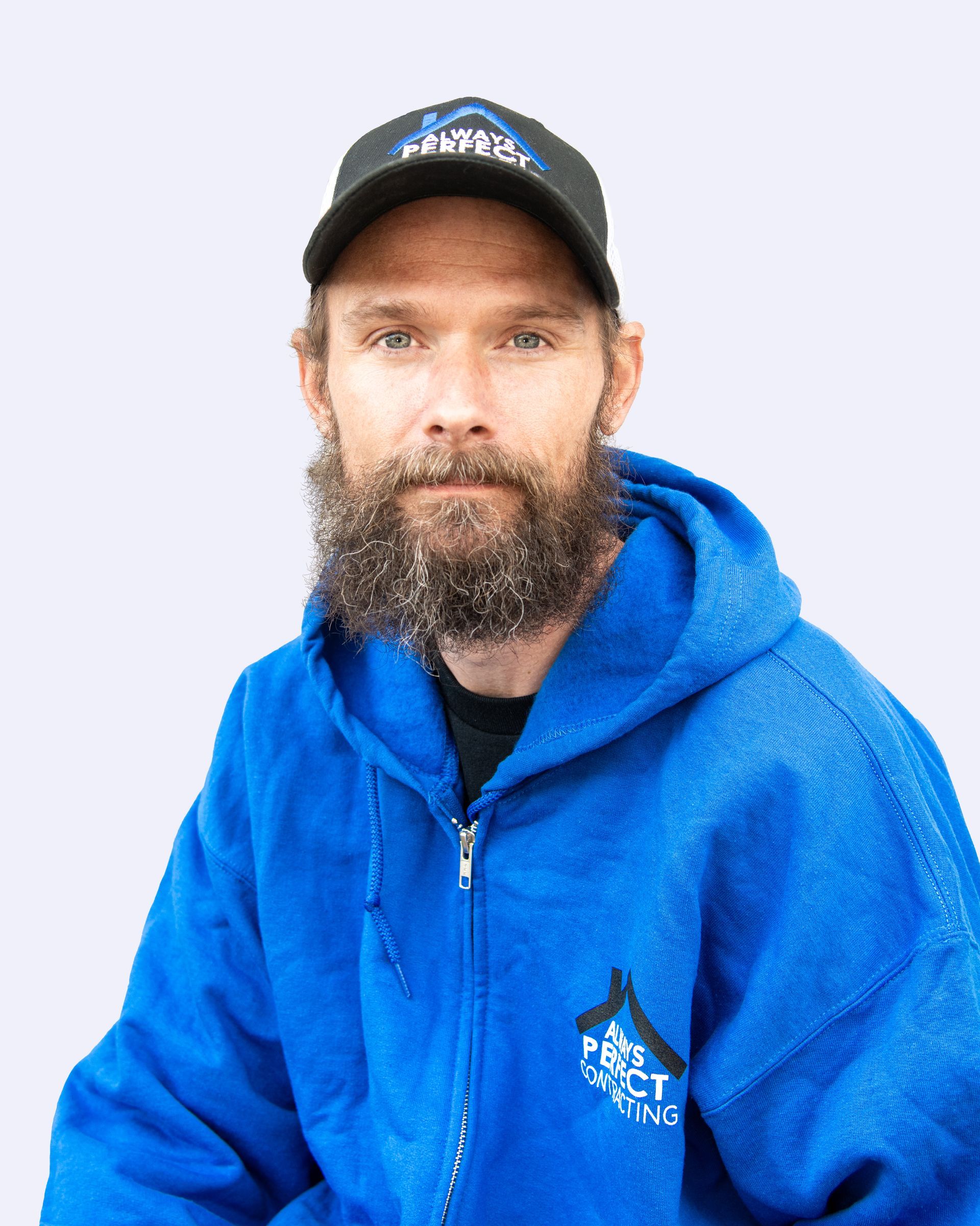 A person with a beard, wearing a black cap and a bright blue hooded zip-up sweatshirt, against a light purple background.