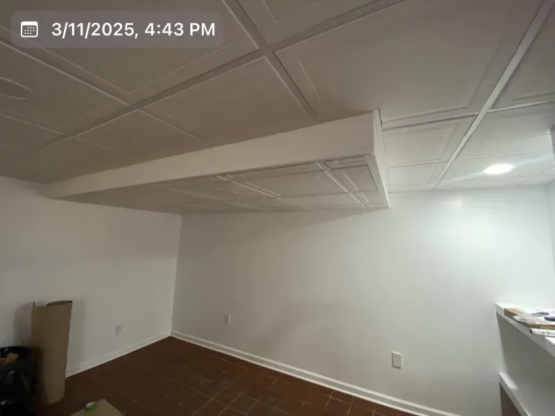 White-walled room with patterned ceiling tiles, brown floor, and a rectangular, protruding white structure.