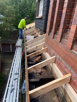 Construction worker on roof with wood supports, adjacent to brick wall.