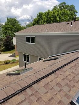 Brown shingled roof with a skylight, grey siding on a building, and trees in the background under a cloudy sky.