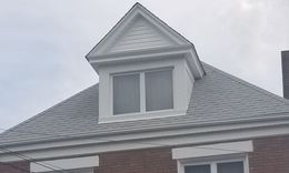 White dormer with two windows on a gray-shingled roof against a cloudy sky.