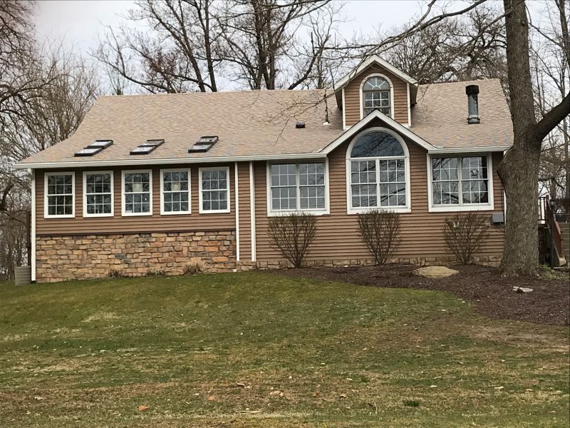 Brown house with beige roof, small dormer, windows, and partial stone foundation, set on a grassy hill.