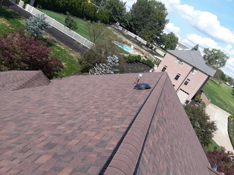 Brown asphalt shingle roof on a house, with a curved ridge cap. Green yard and trees surround.