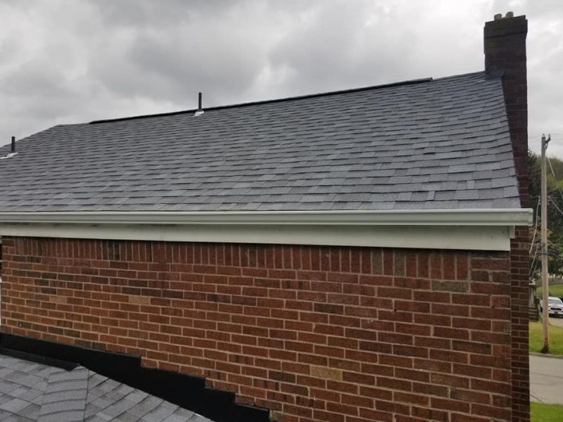 Brick building with a dark gray shingled roof, white gutters, and a brick chimney under a cloudy sky.