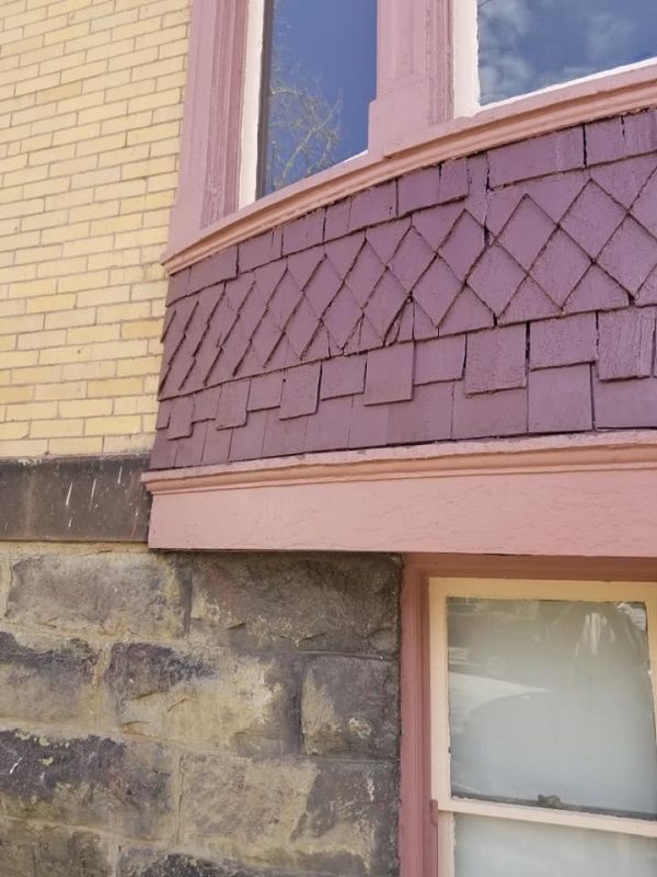 Corner of a building with brick, stone, and wood siding in various colors, including a bay window with decorative shingles.