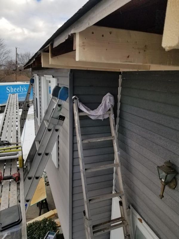 Ladders leaning against a building with gray siding, a wooden overhang, and construction in progress.