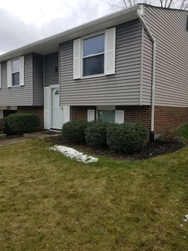 Gray and brick two-story building with white shutters, door, and lawn. Bushes in front and a patch of snow.