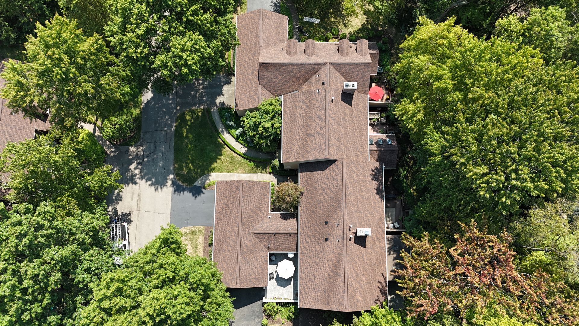 View of a gray shingle roof, chimney vent pipes, and rooftops surrounded by green trees.