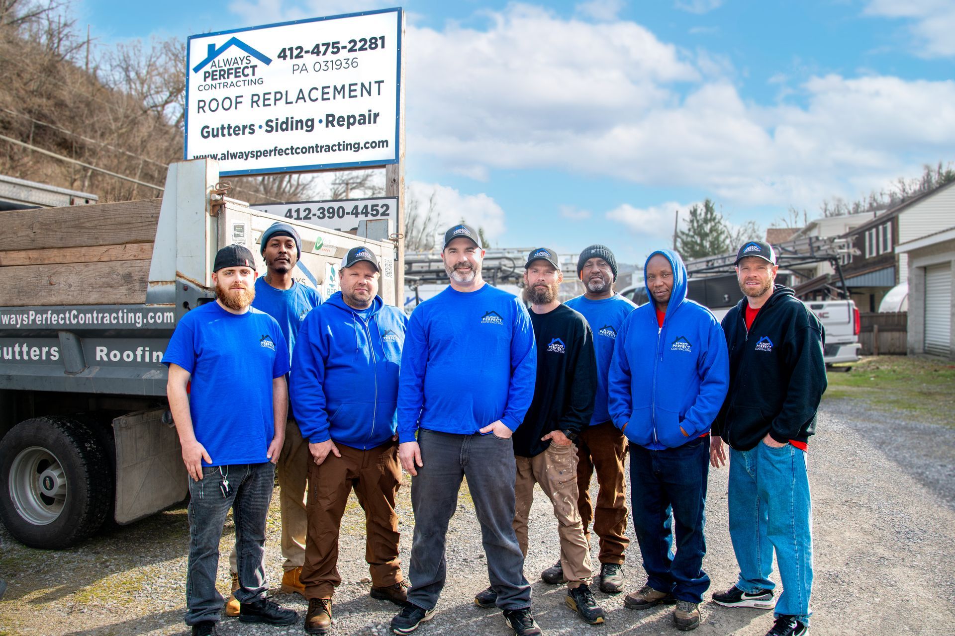 Eight workers in matching blue uniforms stand together in front of a construction truck with a roof replacement sign.