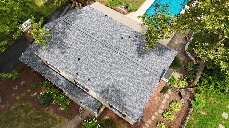 Brown asphalt shingle roof, chimney, and vent, with a green yard and brick house in the background.