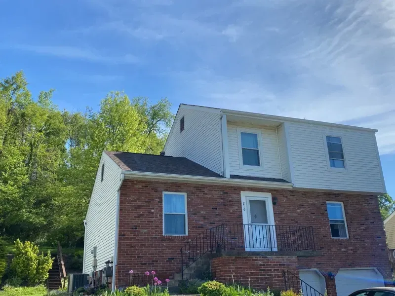 Two-story brick and white-sided house with a dark roof against a blue sky, trees in the background.