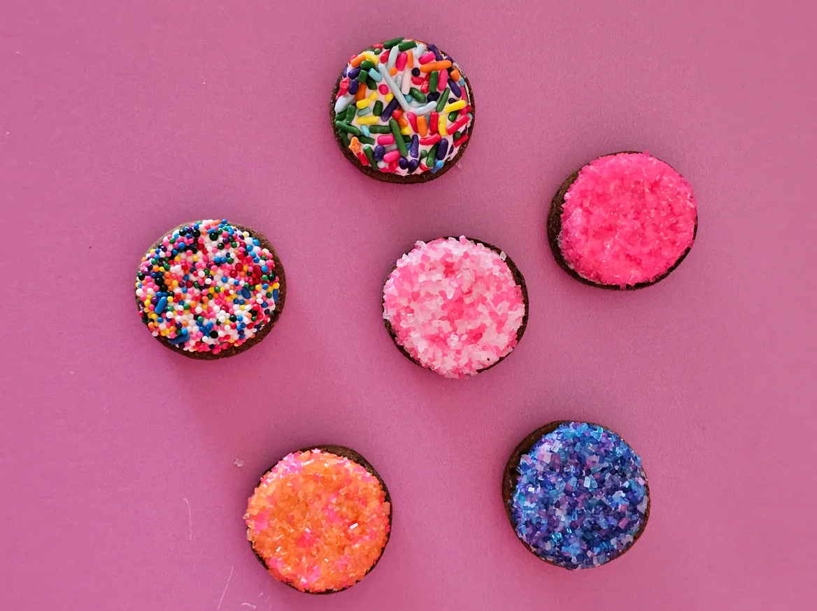 Six cupcakes with different colored frosting and sprinkles on a pink surface.