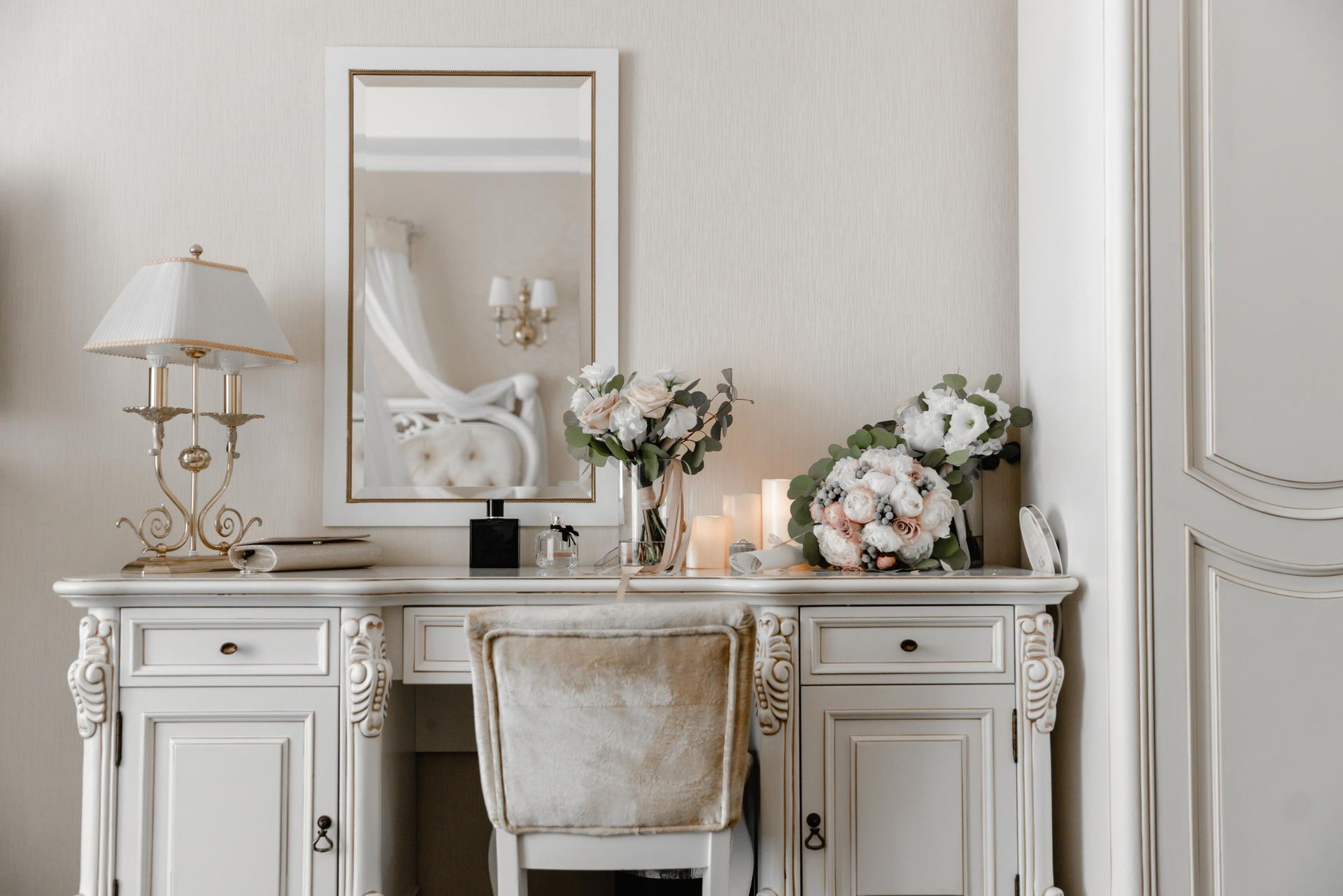 White vanity with a mirror, flowers, and a lamp. 