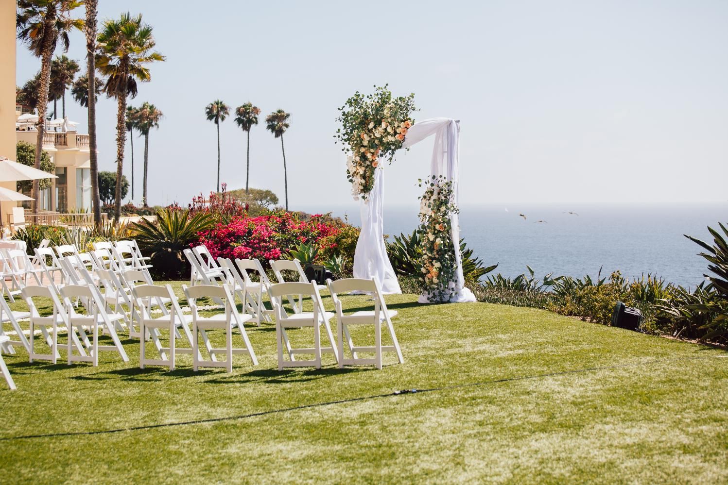 Wedding ceremony setup on a lawn overlooking the ocean: white chairs, floral arch, blue water, and palm trees.