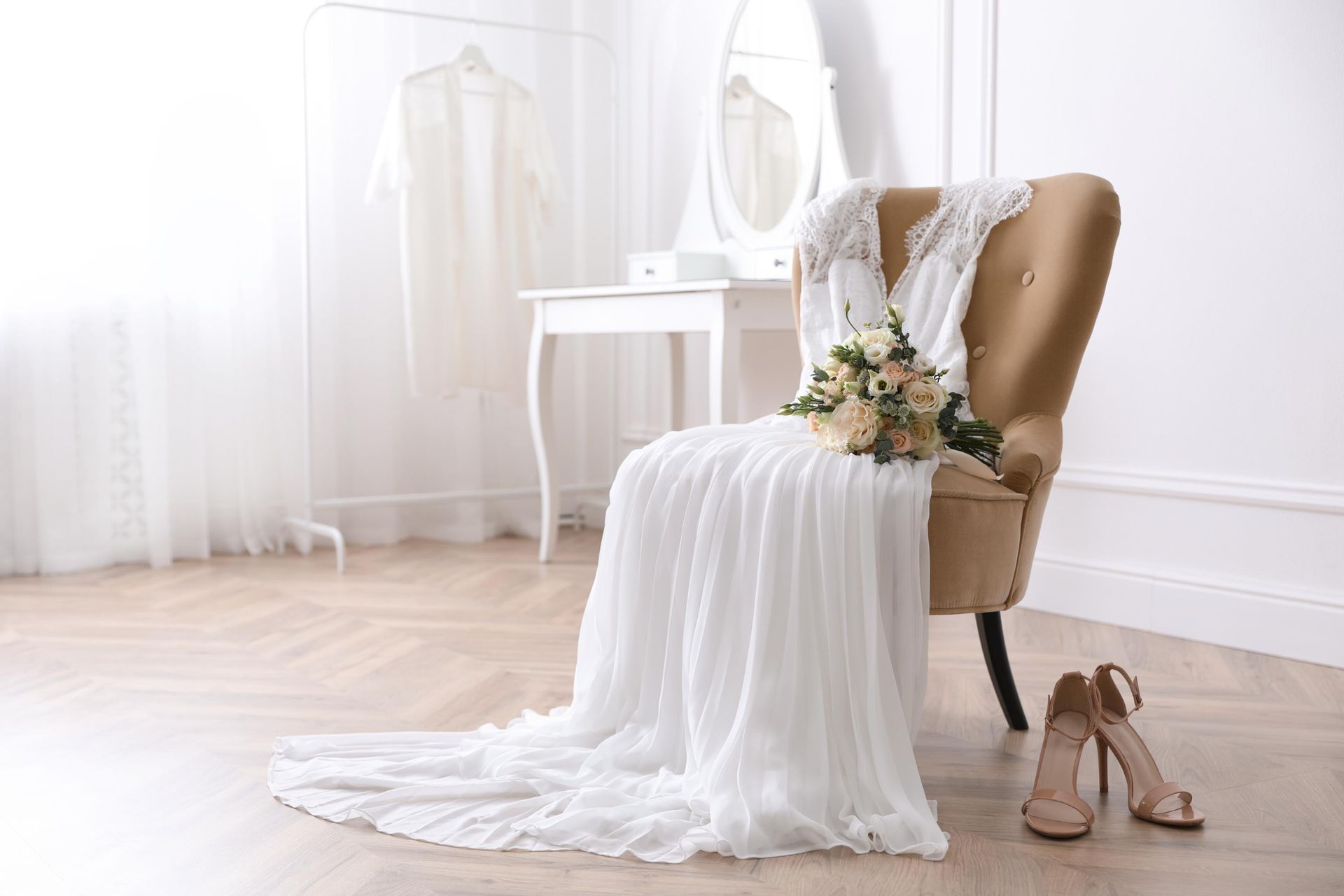 Wedding dress draped on chair next to bridal bouquet and high heels in a light-filled room.