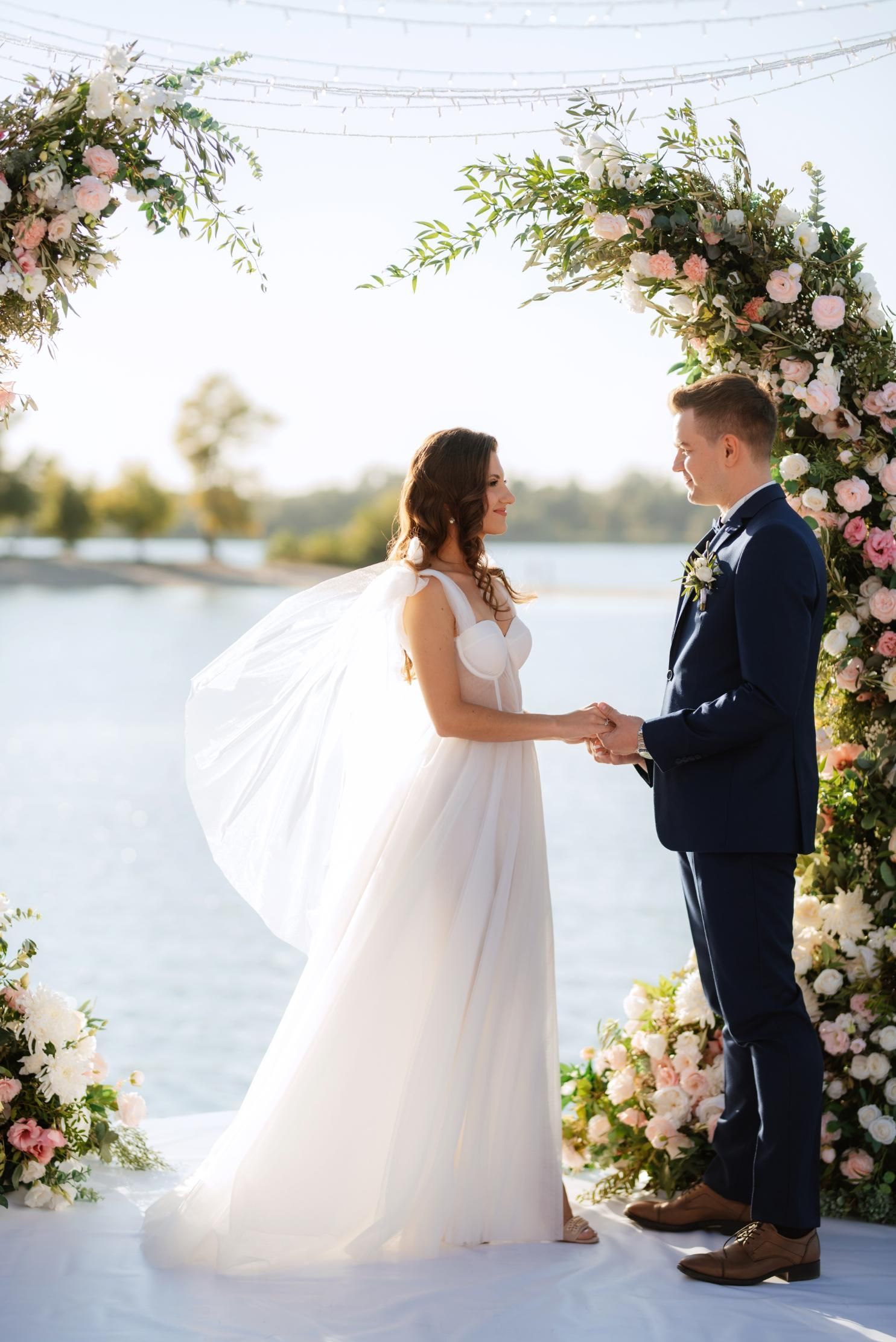 Couple holding hands at wedding ceremony under floral arch. Waterfront setting.