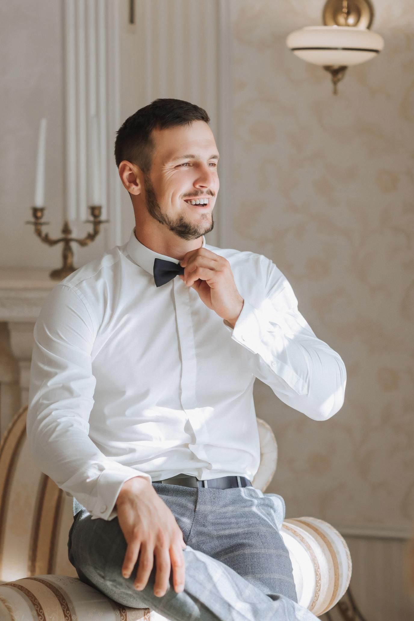 Man in white shirt and bow tie adjusting his collar, seated by a window.