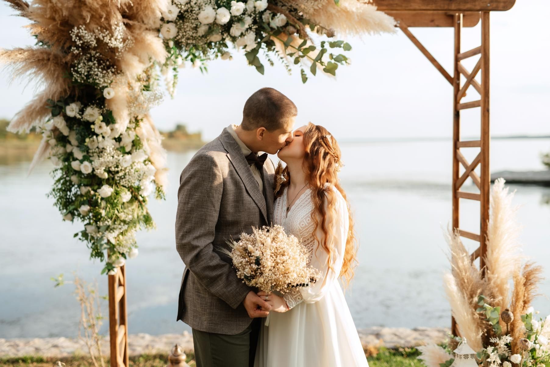 Couple kissing under a floral arch during an outdoor wedding ceremony, with water in the background.