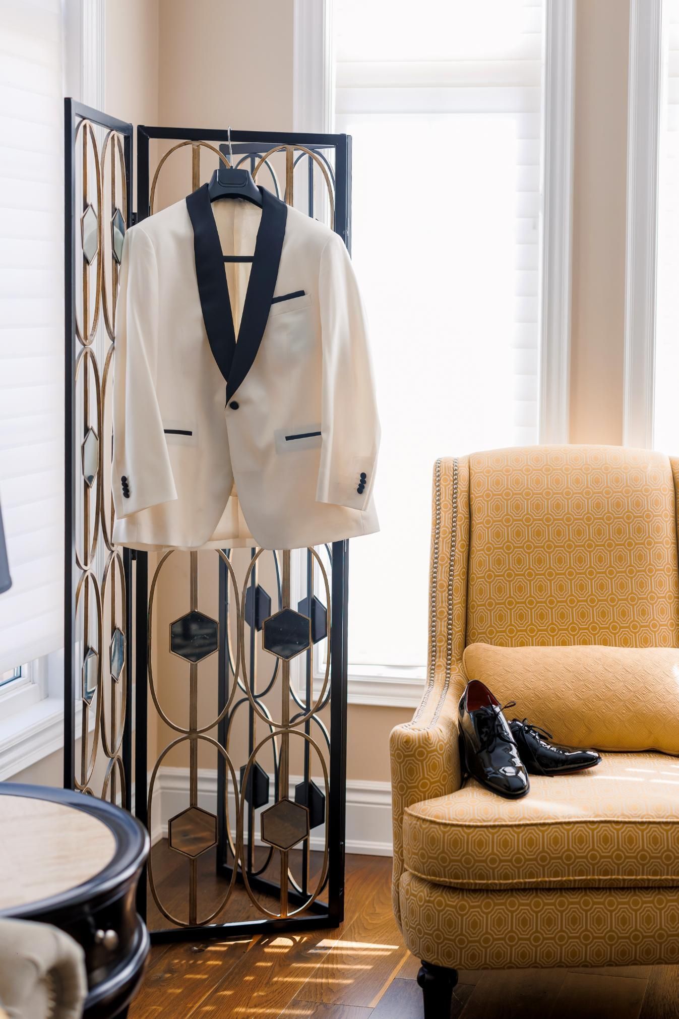 Ivory tuxedo jacket with black lapel hangs on a decorative room divider next to a yellow armchair and dress shoes.