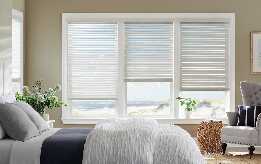 Coastal-themed bedroom with bed, accent chair, and three sets of white horizontal blinds covering the windows.