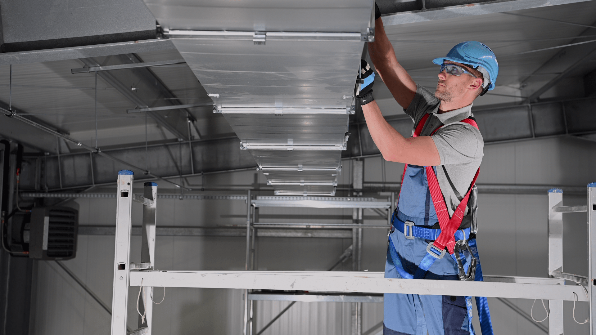 Man in safety gear on a ladder installing ventilation ductwork in a warehouse ceiling.