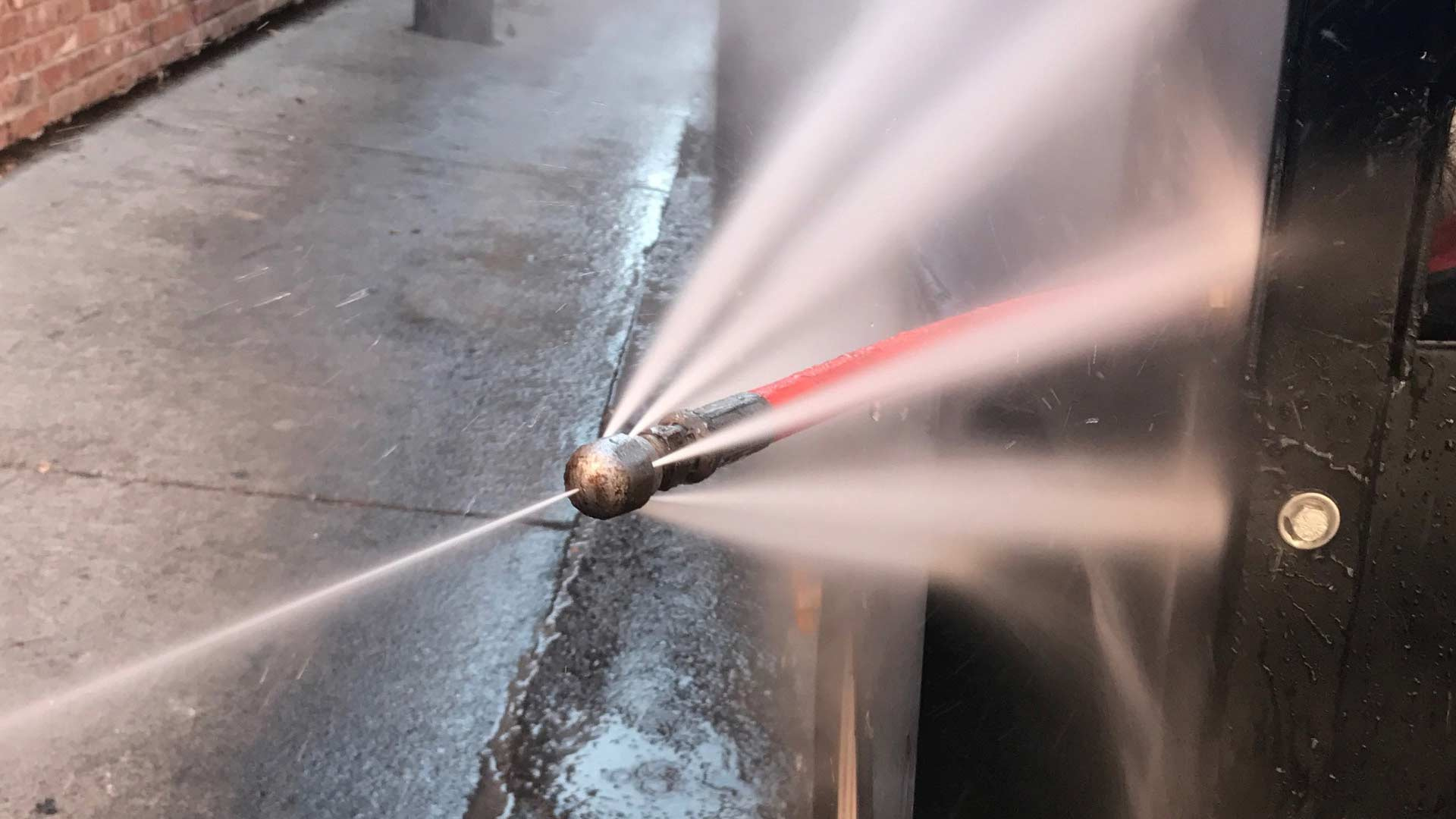 A drain cleaning nozzle sprays water forcefully to clear a drain near a brick wall.