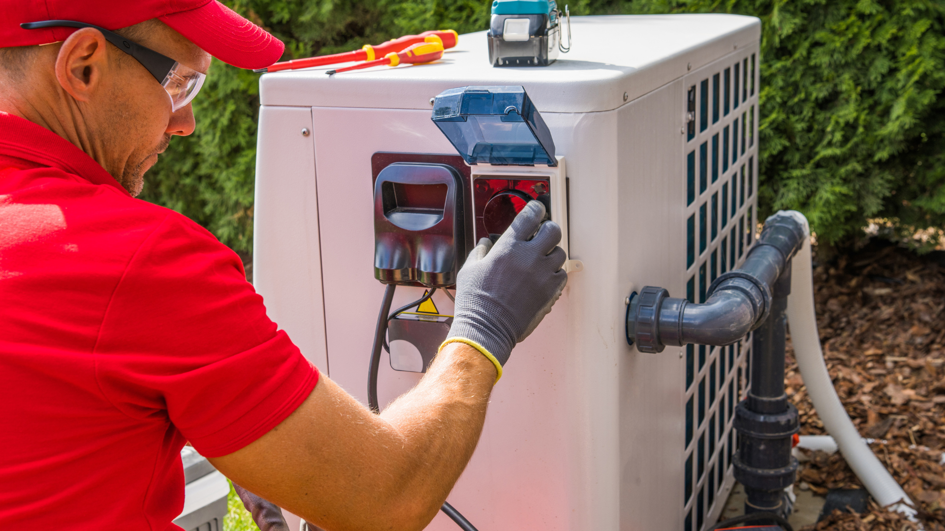 Man in red shirt and gloves working on an outdoor electrical box of a heat pump.