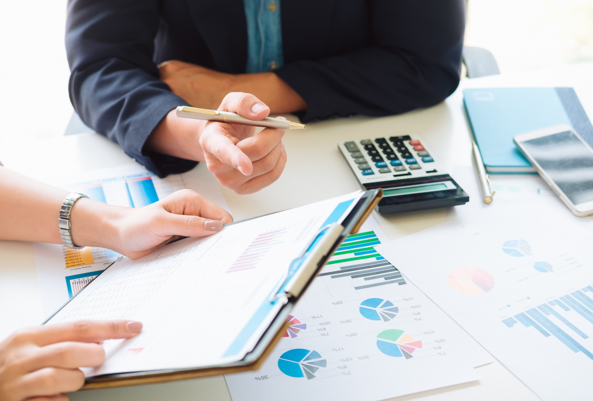 Two people at a desk review charts, one pointing with a pen, with a calculator and phone.