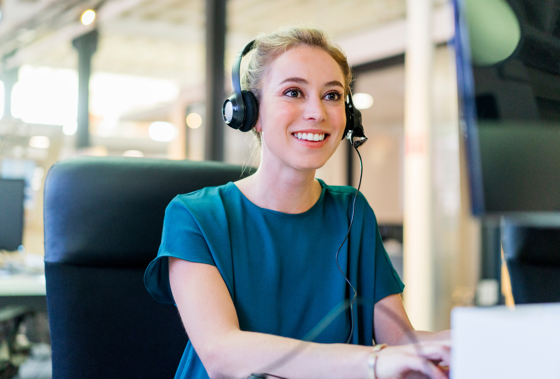 Woman with blonde hair and headset smiles while working at a computer in an office.
