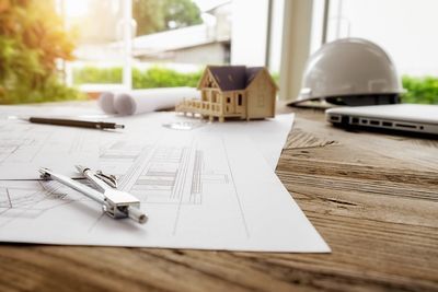 A wooden table topped with drawings , a compass , and a model house.