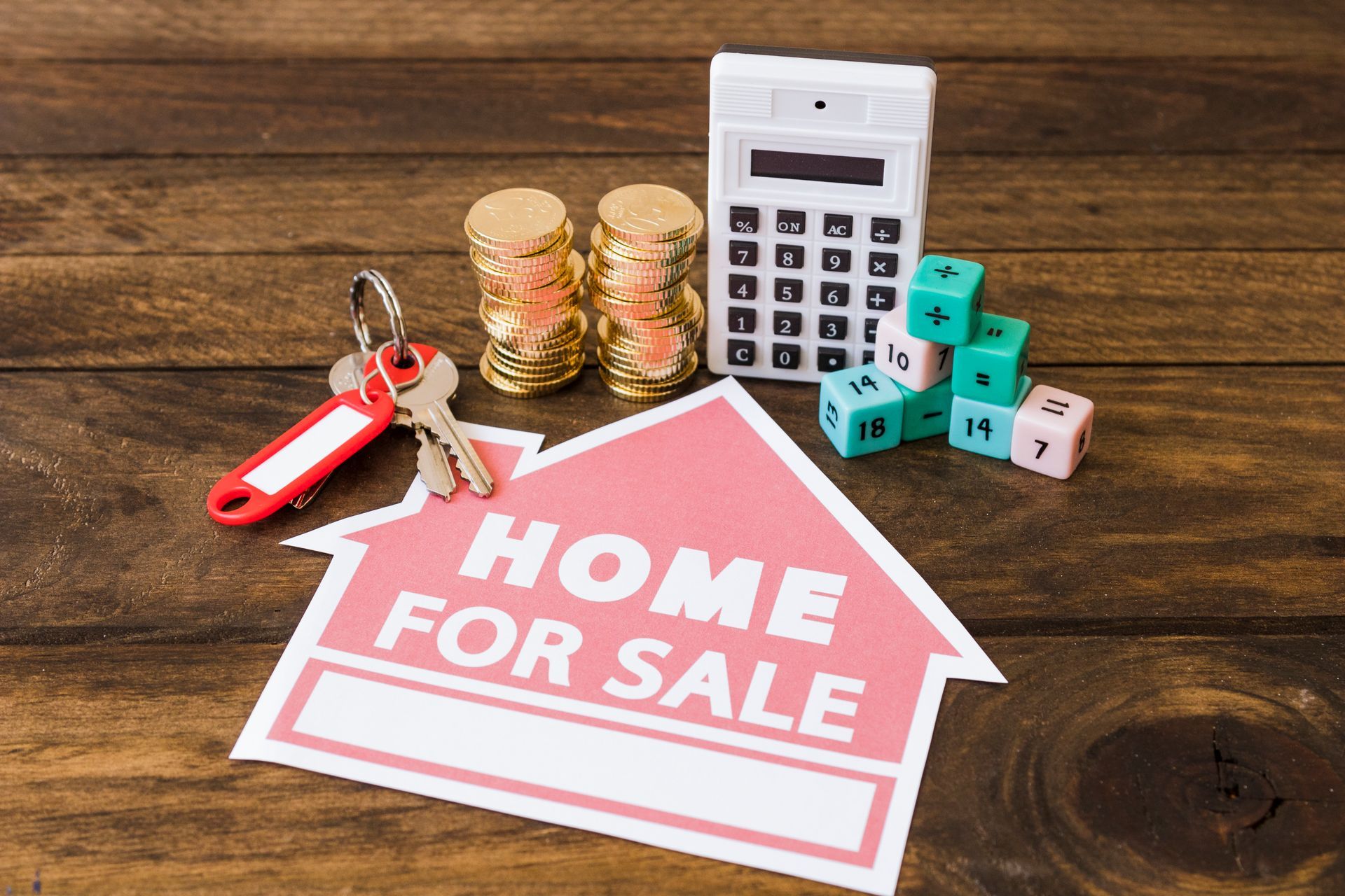 A home for sale sign , keys , coins , dice and a calculator on a wooden table.