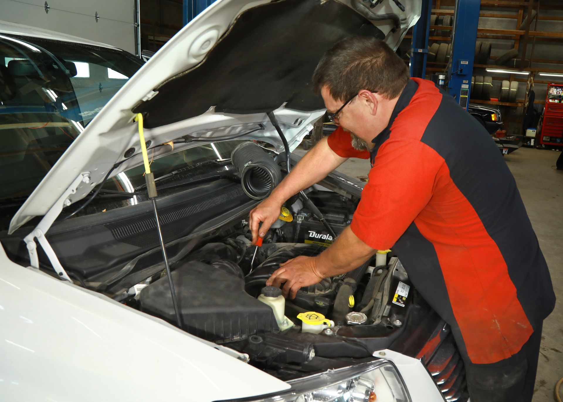 Mechanic working on the engine of a white car in a garage, wearing a red and black shirt.