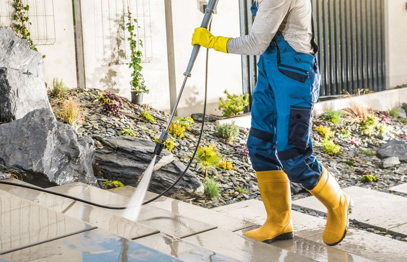 Worker in yellow boots pressure-washes a sidewalk beside rocks and plants.