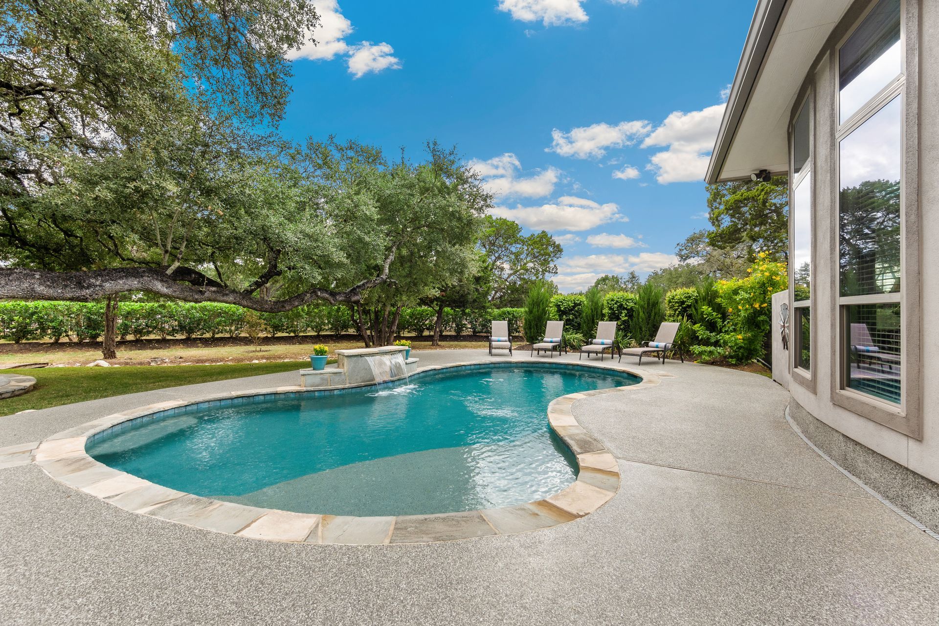 Backyard pool with curved stone deck, lounge chairs, and lush trees under a blue sky