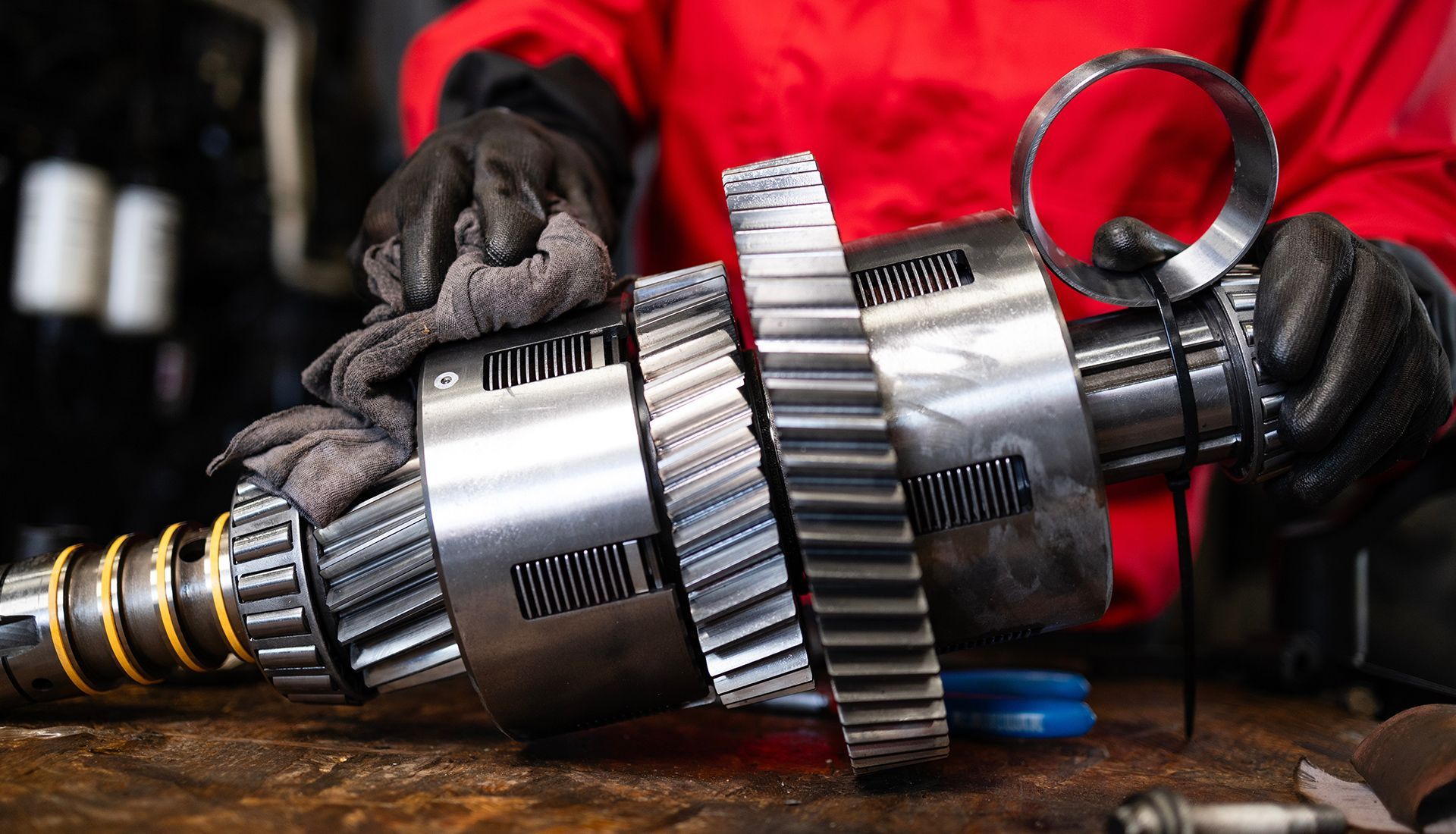 A mechanic in red overalls and gloves working on disassembled gears, holding a metal ring.
