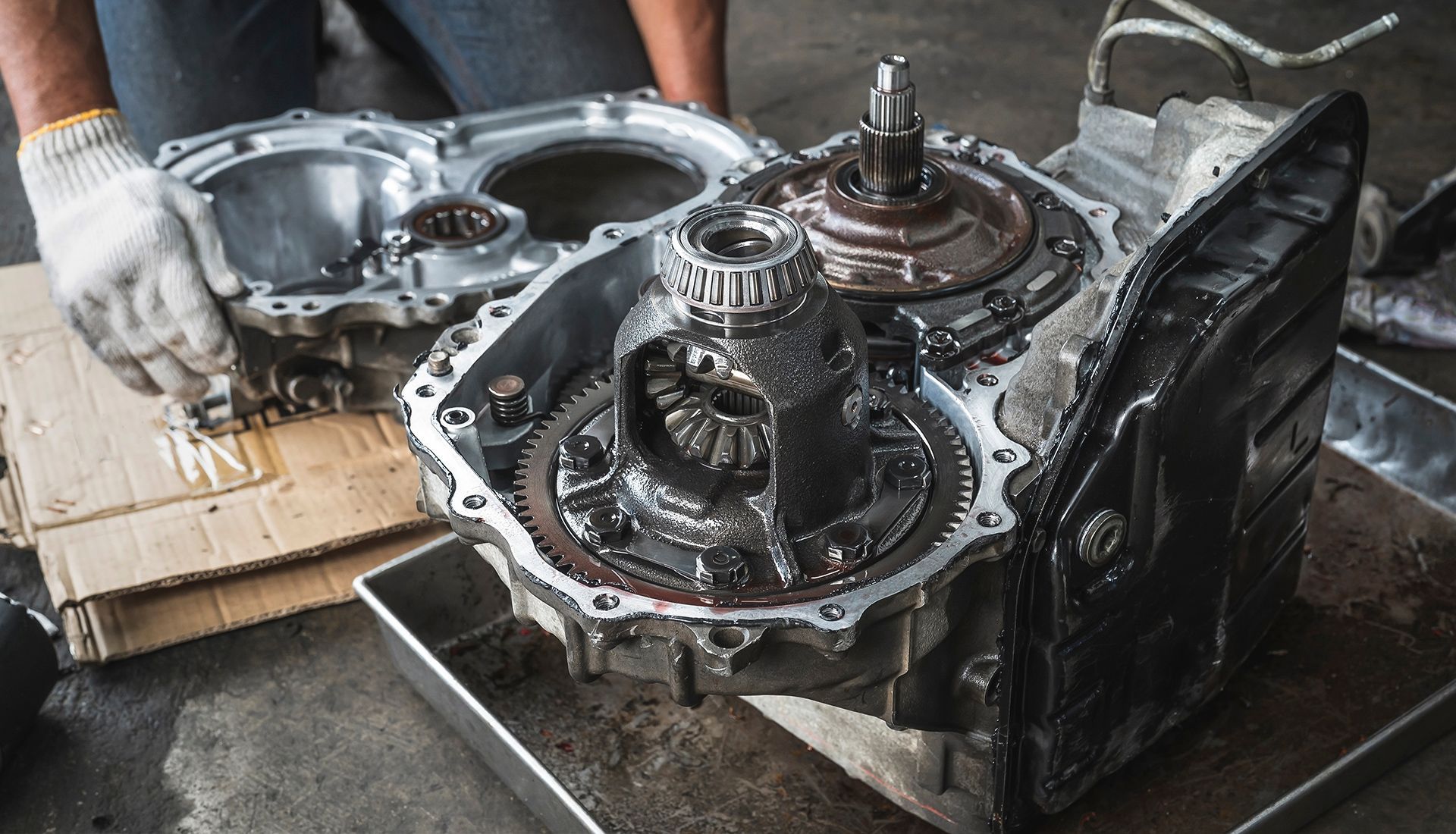 A mechanic's gloved hand working on an open car transmission, showing gears, housing, and components in a shop setting.