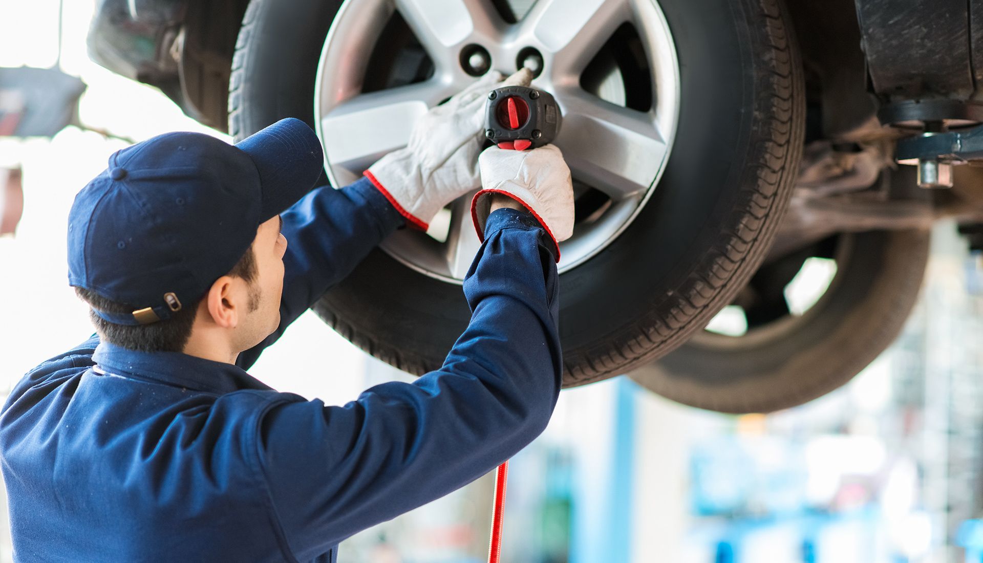 Mechanic in blue coveralls and cap uses a power tool to work on a car tire raised on a lift in a repair shop.