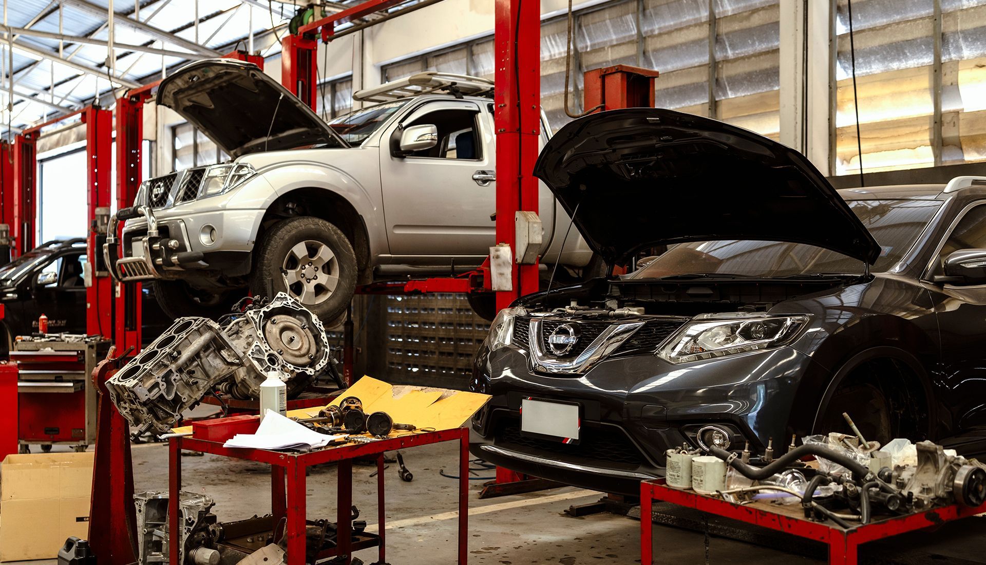 Cars being repaired in an auto shop. A silver truck is lifted, a black car has its hood open. Parts are on red tables.