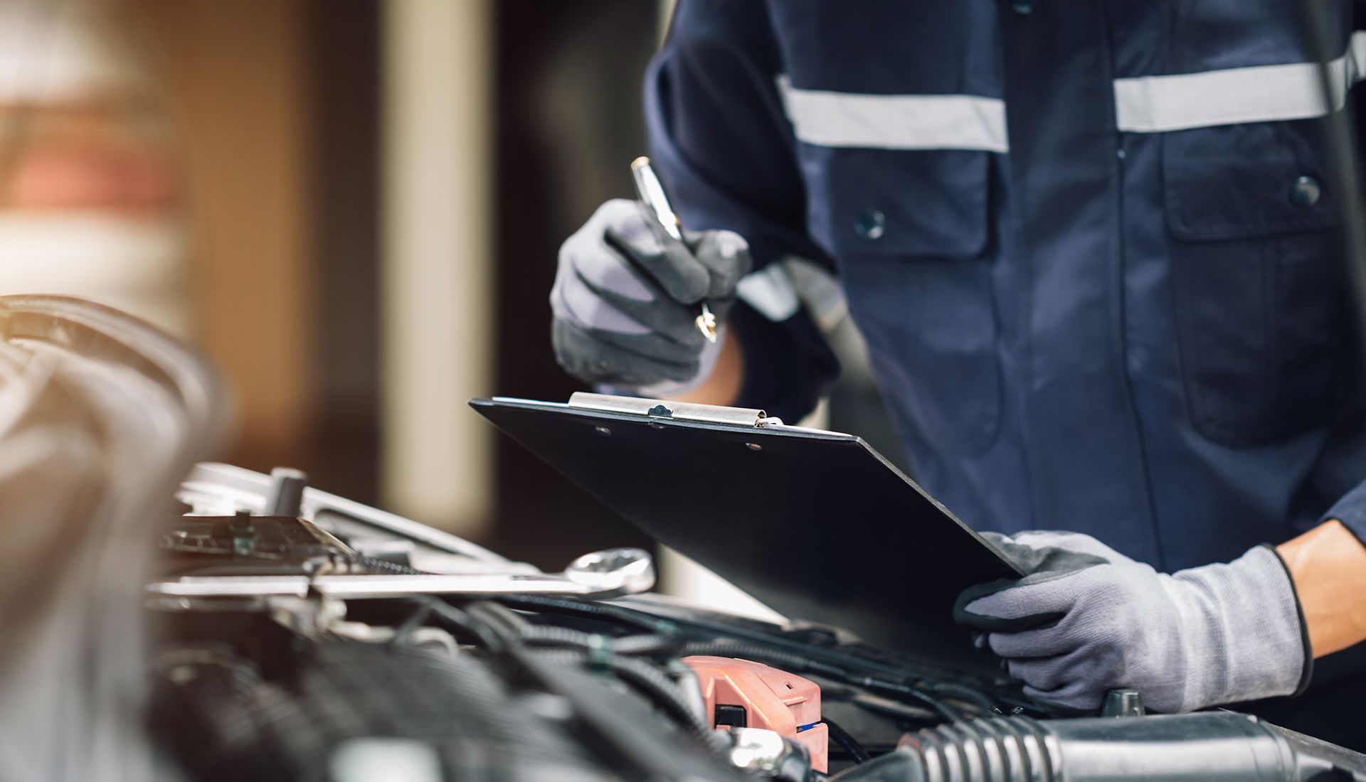 Mechanic in blue work suit, wearing gloves, inspecting a car engine and writing on a clipboard.