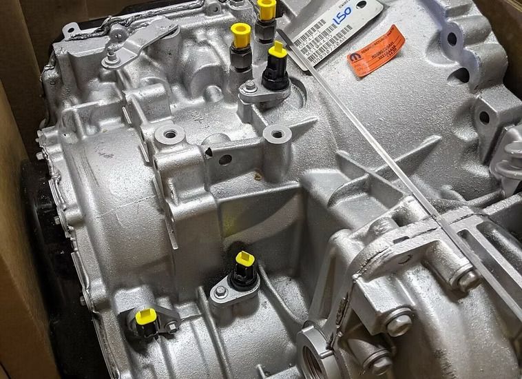New silver automatic transmission in a cardboard box. Various yellow and black caps cover the openings.